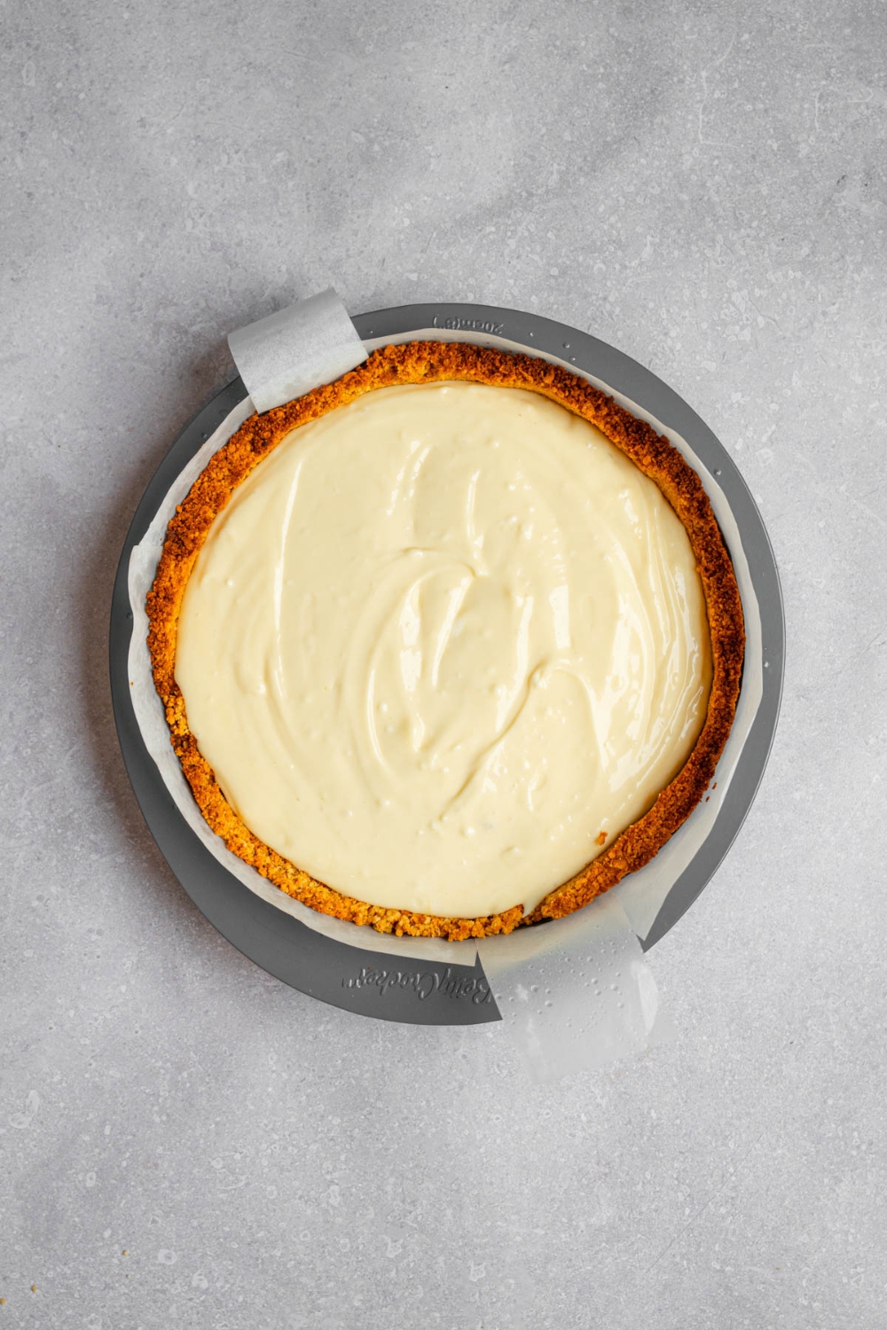 A cheesecake pan with uncooked creme brulee cheesecake in a graham cracker crumb crust. The pan is on a white counter.
