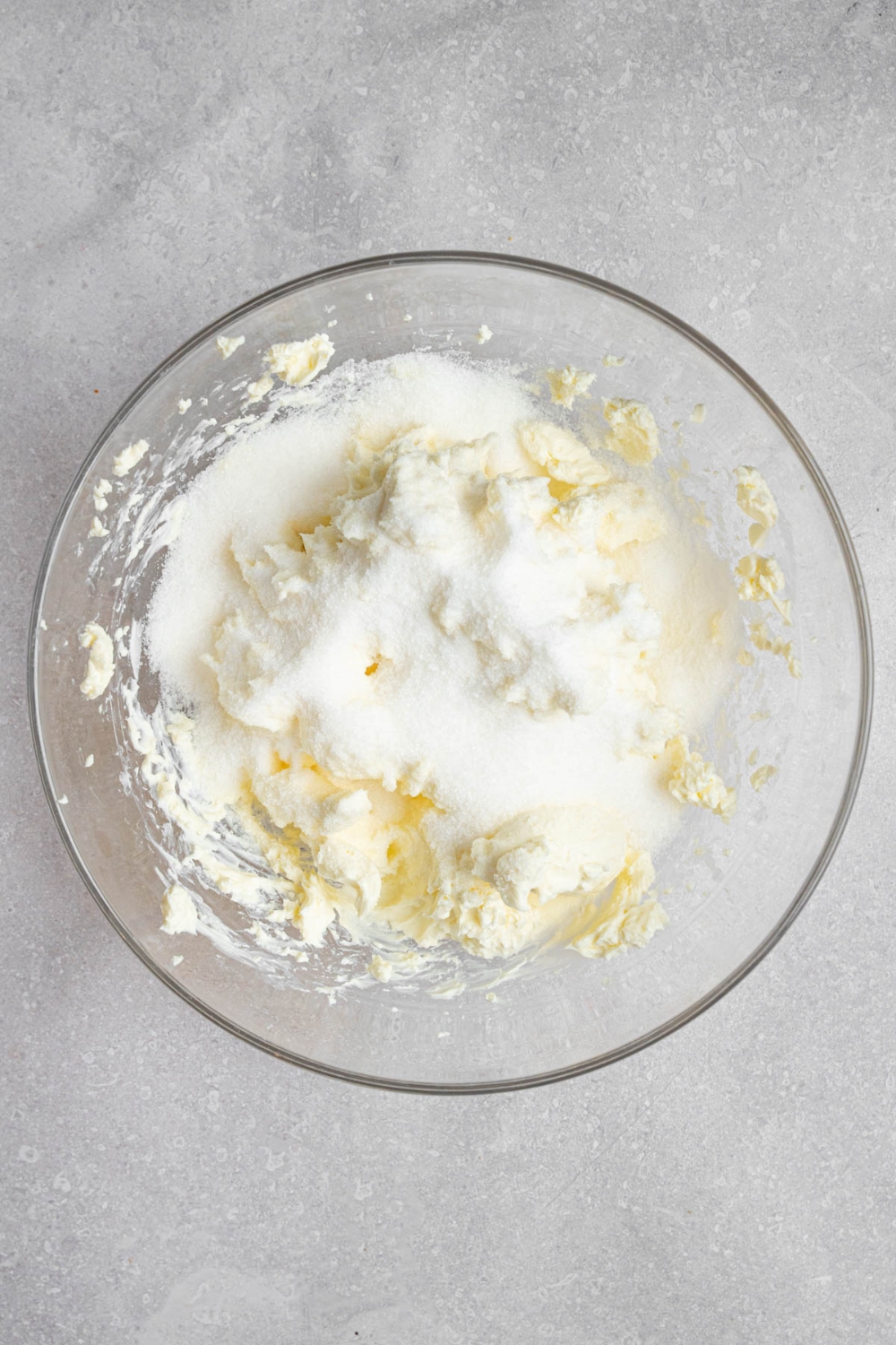 A glass bowl with beaten cream cheese and sugar. The bowl is on a white counter.