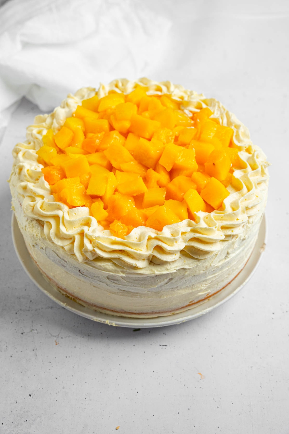 A whole mango cake topped with cubed mango served on a white plate. The plate is on a white counter with a white cloth napkin.