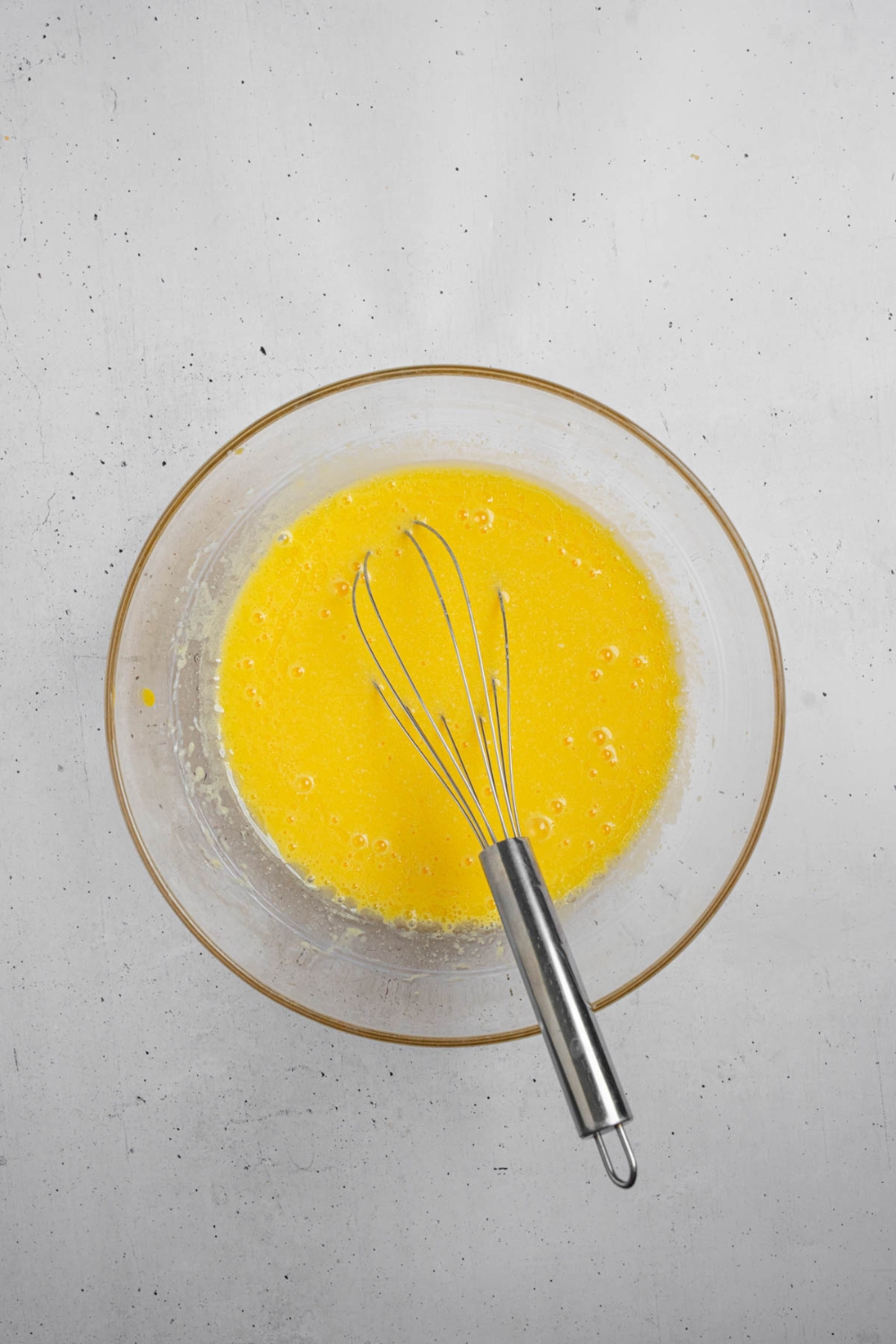 A glass bowl with a whisk mixing wet ingredients for mango cake. The bowl is on a white counter.