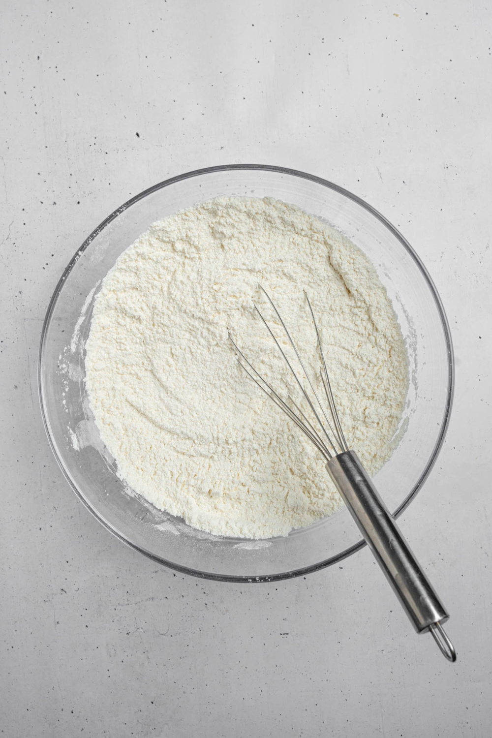 A glass bowl with a whisk mixing dry ingredients for mango cake. The bowl is on a white counter.