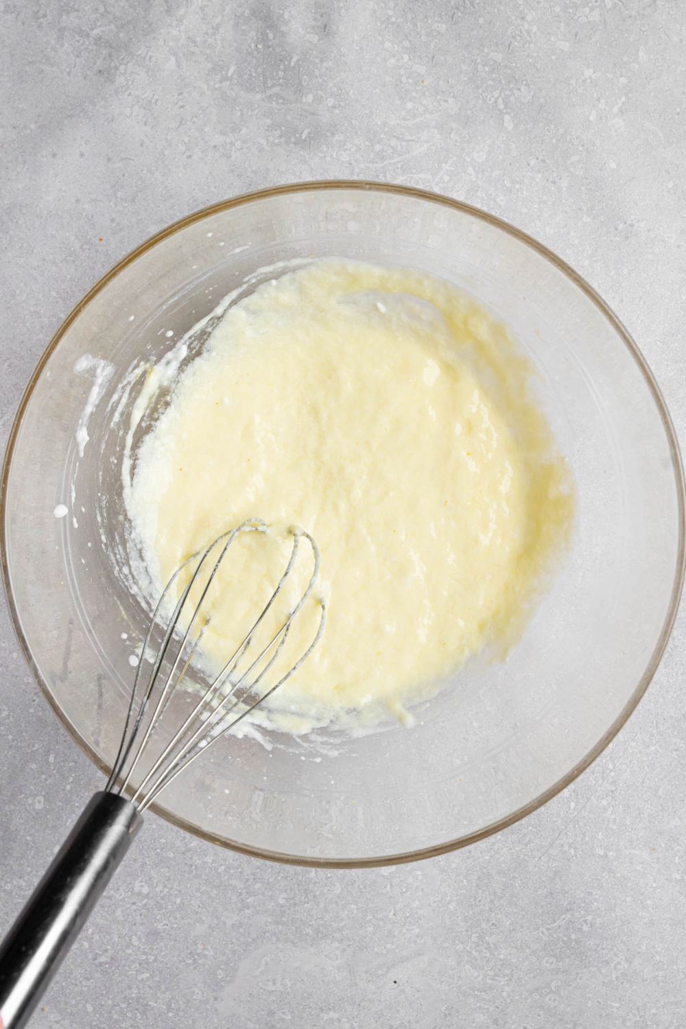 A glass bowl with a whisk mixing wet ingredients for mini pancake batter. The bowl is on a white counter.