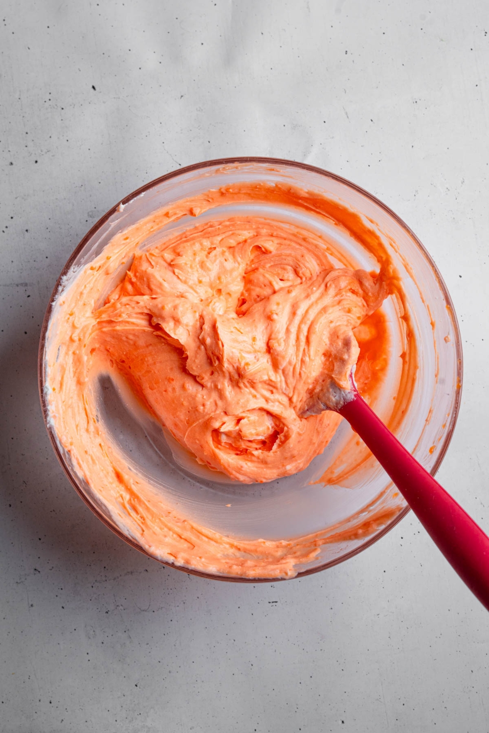 A glass bowl with a spatula stirring a strawberry jello and cool whip mixture. The bowl is on a white counter.