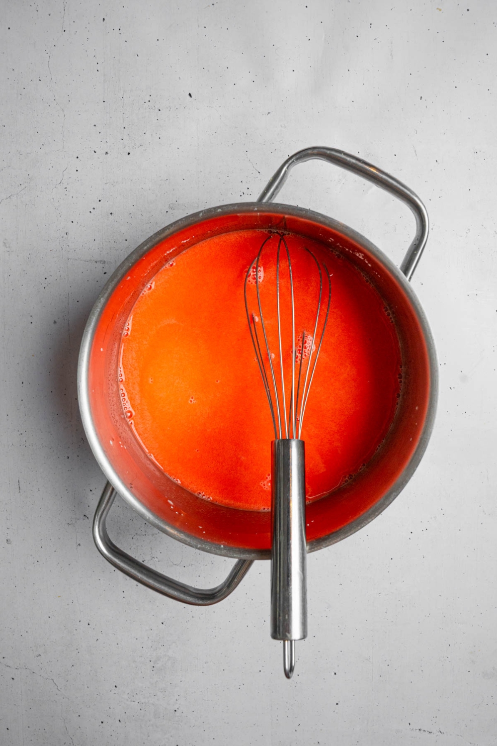 A whisk inside a stock pot with strawberry jello mixture. The pot is on a white counter.