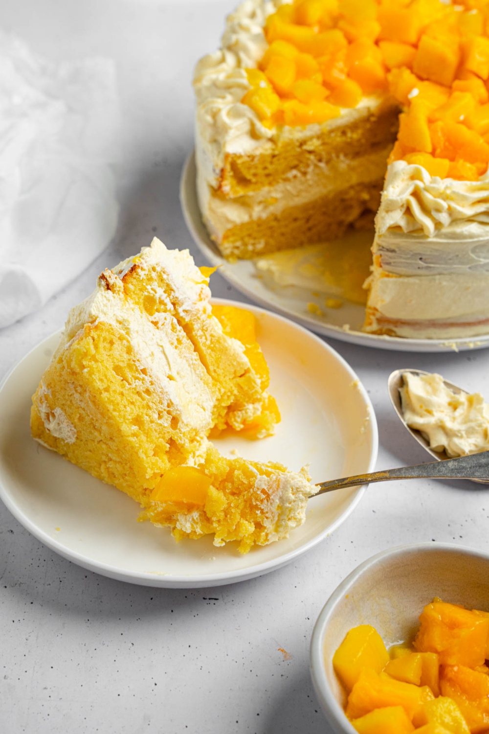 A slice of mango cake on a white plate with a fork taking a bite of cake. The plate is on a white counter with a whole mango cake and bowl of cubed mango.