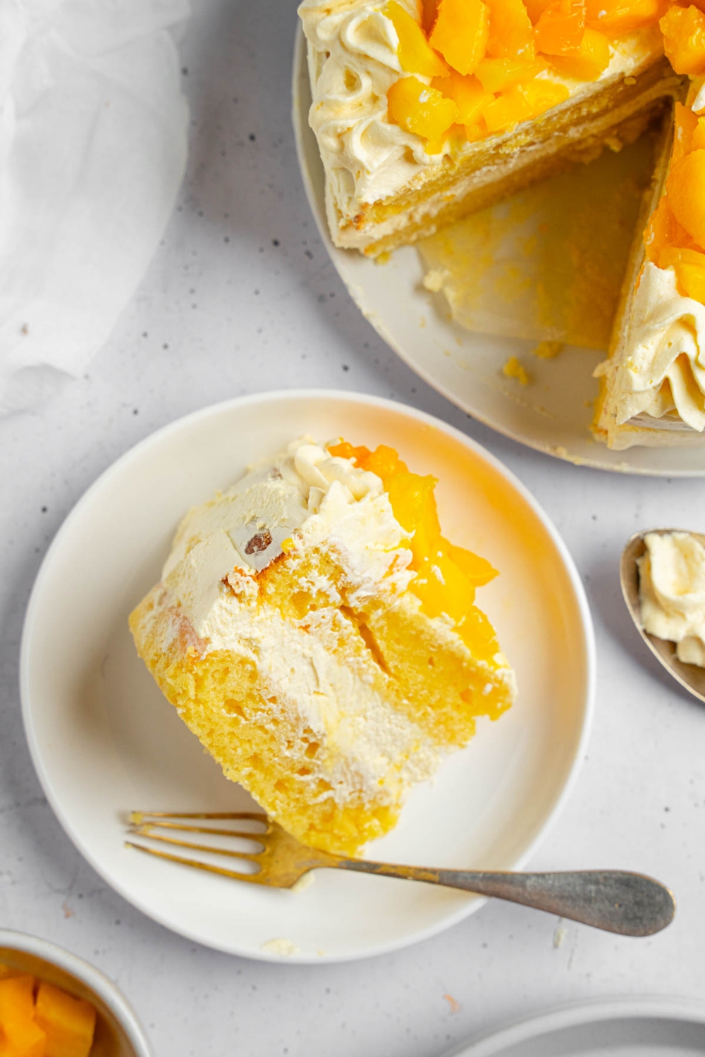A slice of mango cake on a white plate with a fork. The plate is on a white counter with a whole mango cake and bowl of cubed mango.