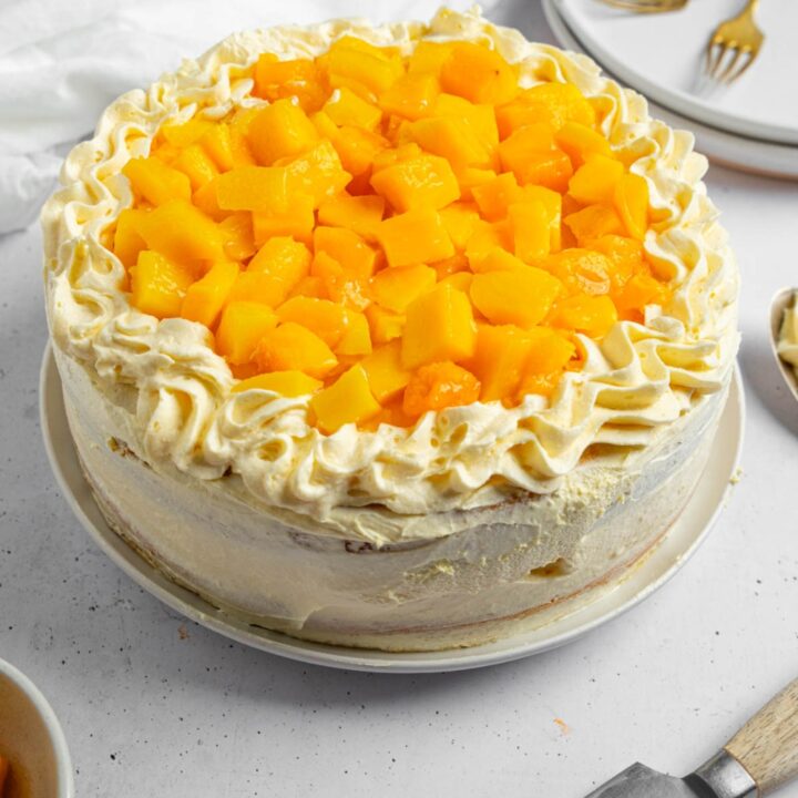 A whole mango cake topped with cubed mango. The cake is on a white counter with a knife, bowl of mango, and plate with two forks.