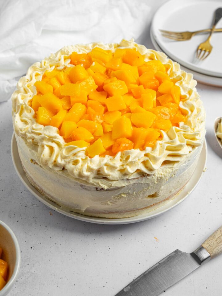 A whole mango cake topped with cubed mango. The cake is on a white counter with a knife, bowl of mango, and plate with two forks.