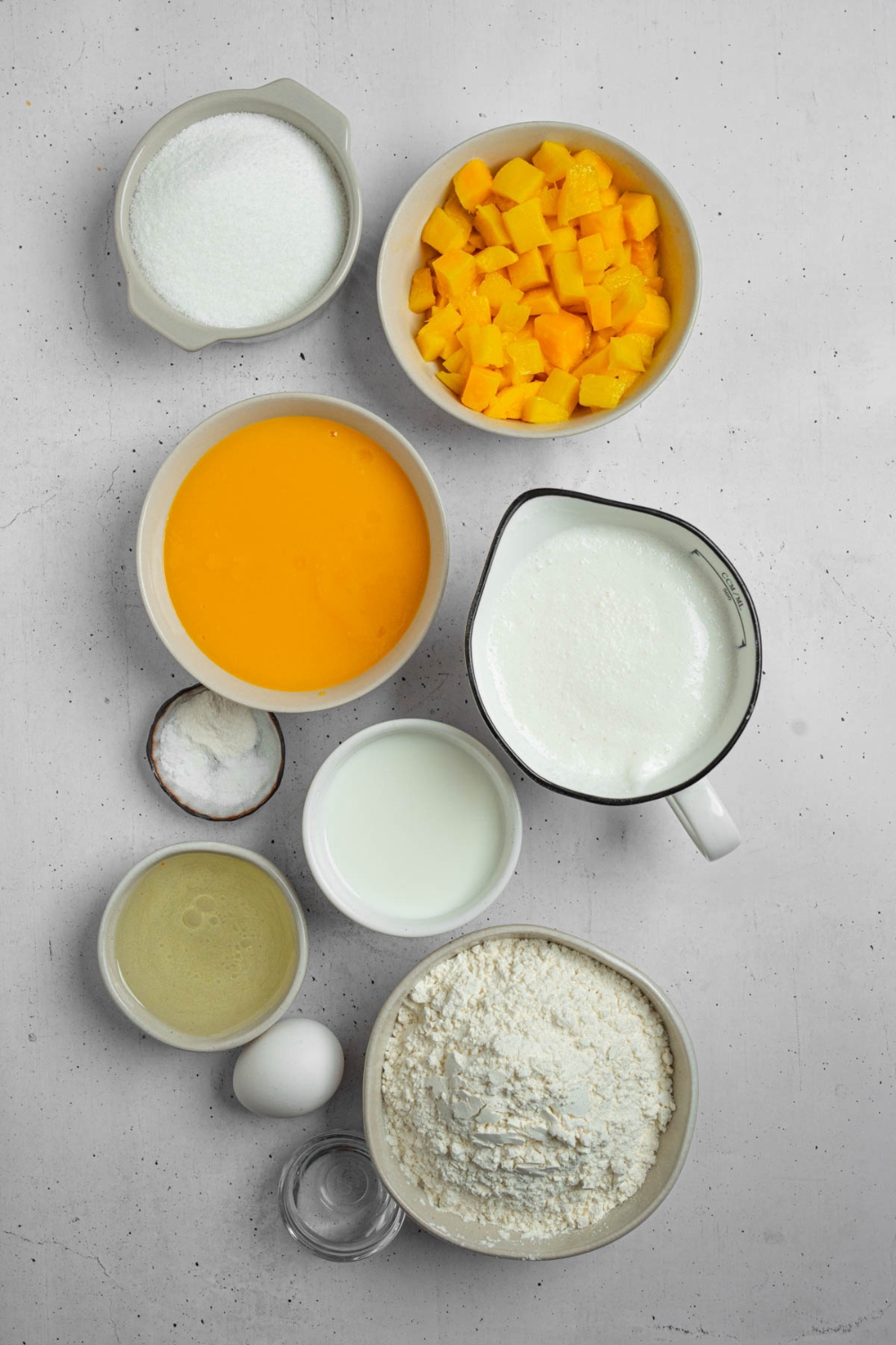 An overhead shot of several bowls in various sizes containing ingredients to make mango cake including mango puree, diced mango, flour, egg, vanilla extract, sugar, baking soda, and more.