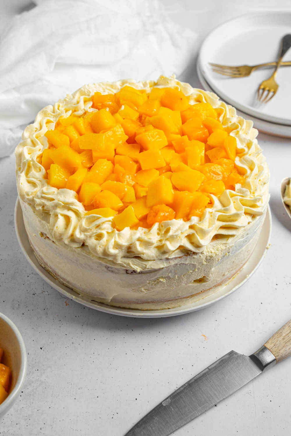 A whole mango cake topped with cubed mango. The cake is on a white counter with a knife, bowl of mango, and plate with two forks.