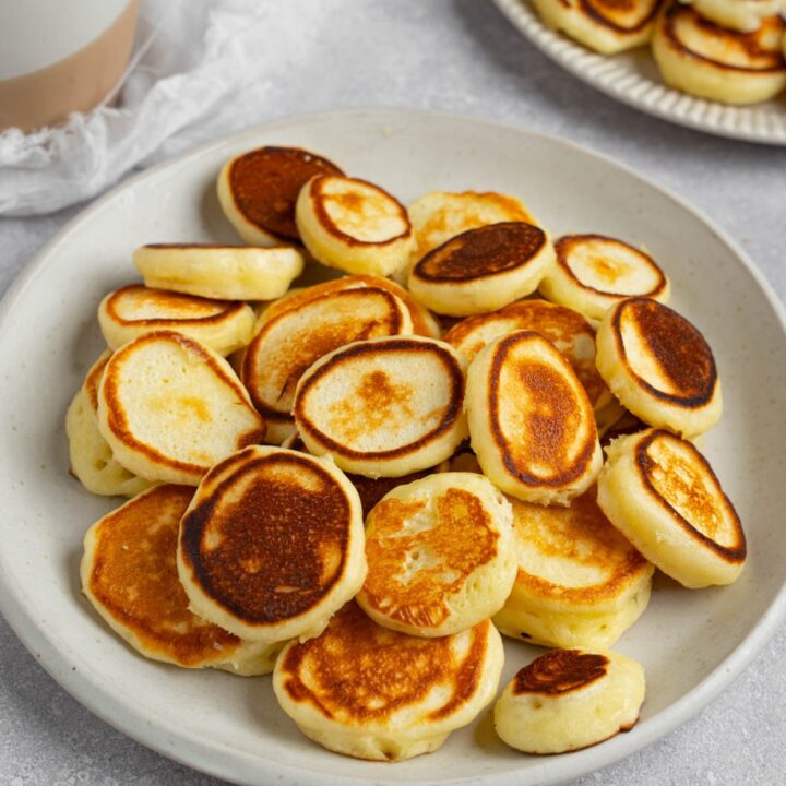 A white plate filled with mini pancakes. The plate is on a white counter with a cup of coffee and additional plate of pancakes.