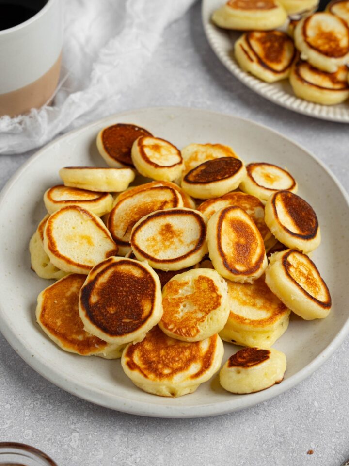 A white plate filled with mini pancakes. The plate is on a white counter with a cup of coffee and additional plate of pancakes.
