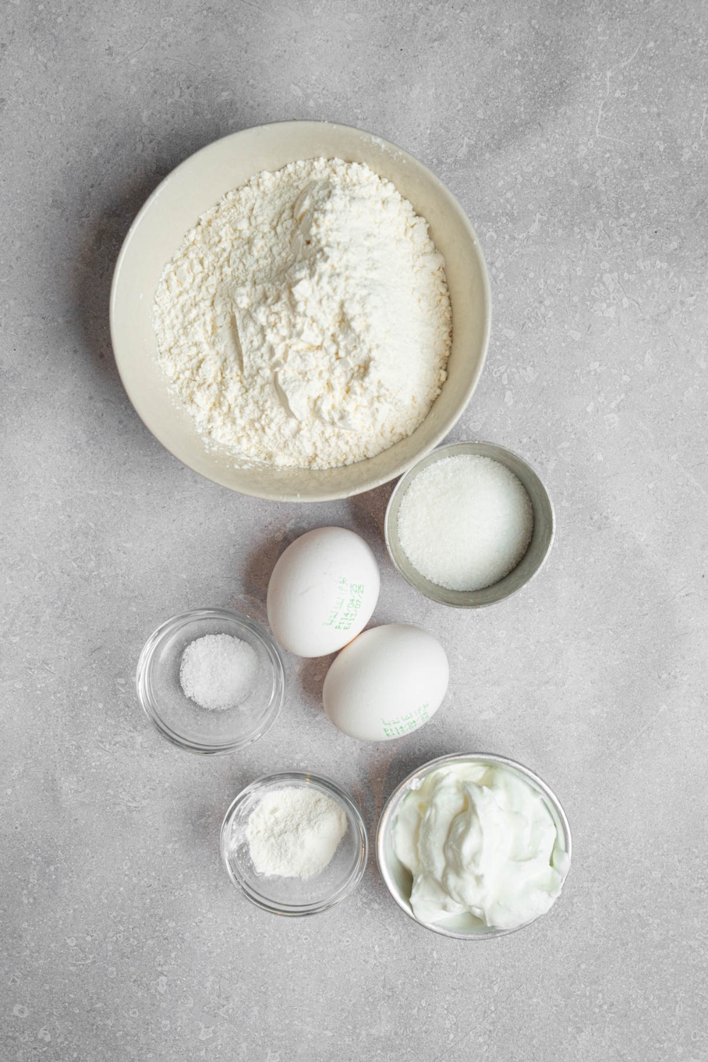 An overhead shot of several bowls in various sizes containing ingredients to make mini pancakes including eggs, sour cream, flour, sugar, salt, and baking powder.