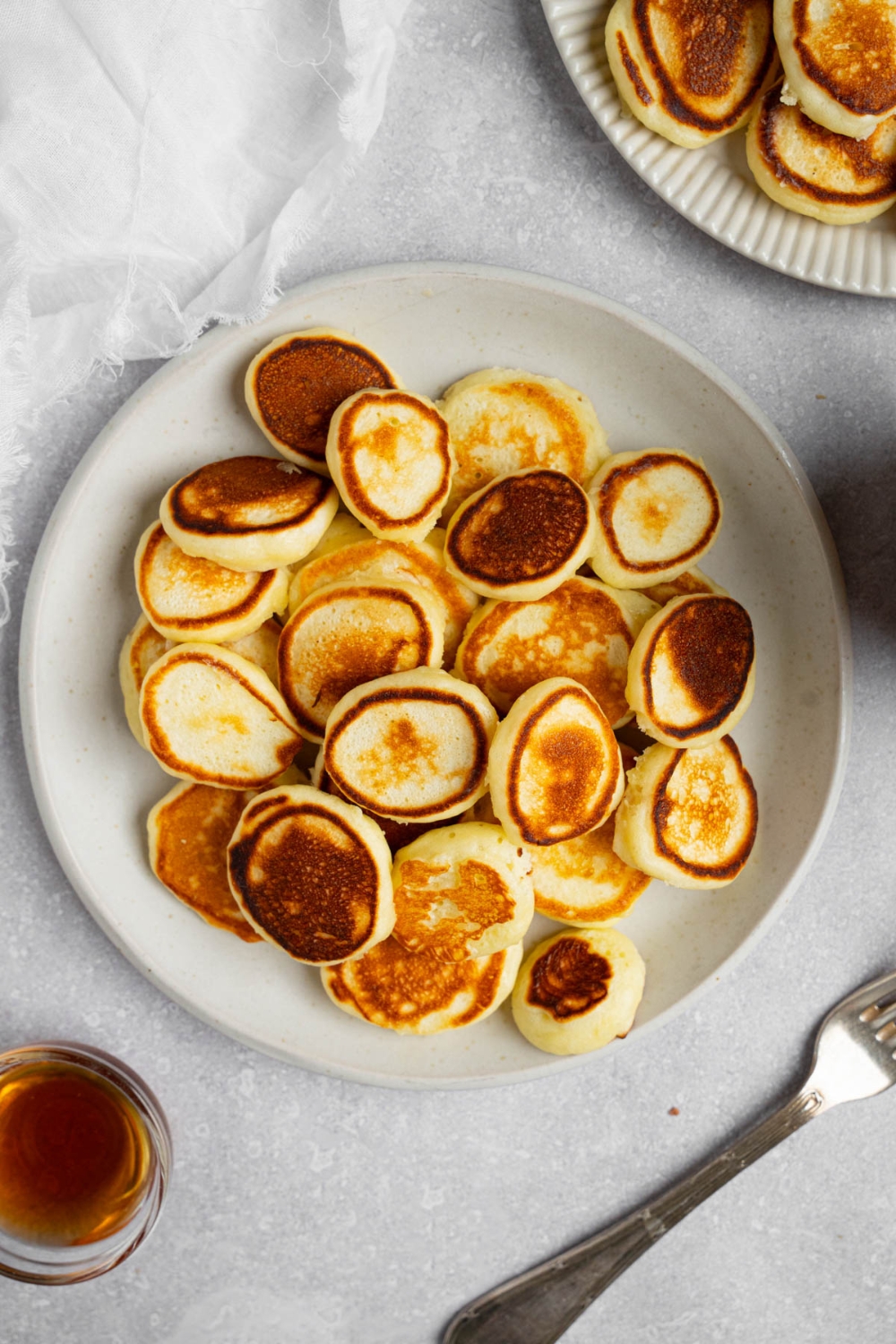 A white plate filled with mini pancakes. The plate is on a white counter with a fork, small glass bowl of syrup, and an additional plate of pancakes.