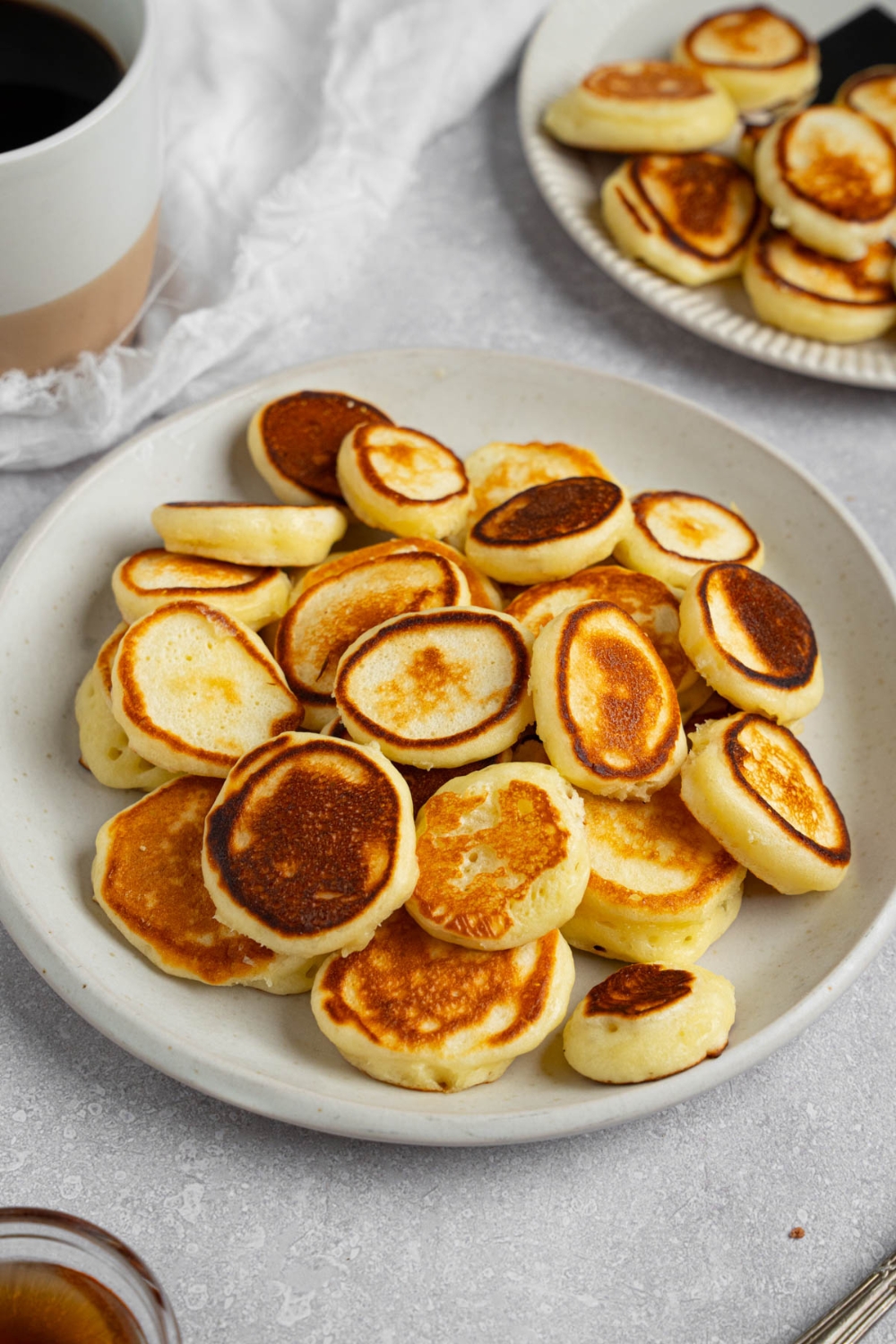 A white plate filled with mini pancakes. The plate is on a white counter with a cup of coffee and additional plate of pancakes.