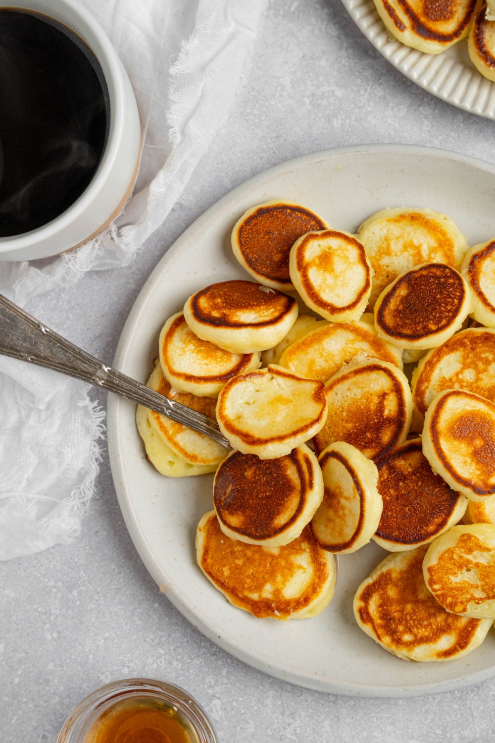 A white plate filled with mini pancakes with a fork underneath them. The plate is on a white counter with a cup of coffee and additional plate of pancakes.