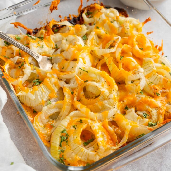 A square glass baking dish with Tennessee onions garnished with fresh parsley. There is a fork in the onions. The dish is on a white counter with a bowl of sour cream, garnishes, and brick of cheese.