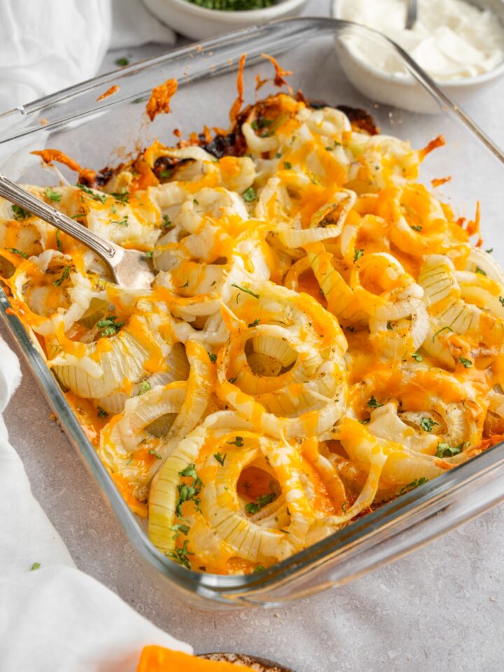 A square glass baking dish with Tennessee onions garnished with fresh parsley. There is a fork in the onions. The dish is on a white counter with a bowl of sour cream, garnishes, and brick of cheese.