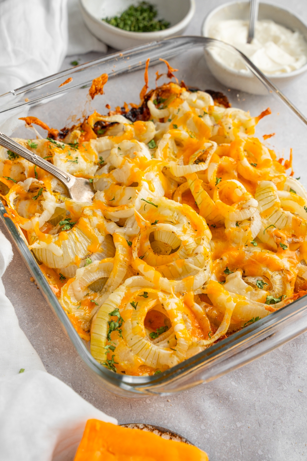 A square glass baking dish with Tennessee onions garnished with fresh parsley. There is a fork in the onions. The dish is on a white counter with a bowl of sour cream, garnishes, and brick of cheese.