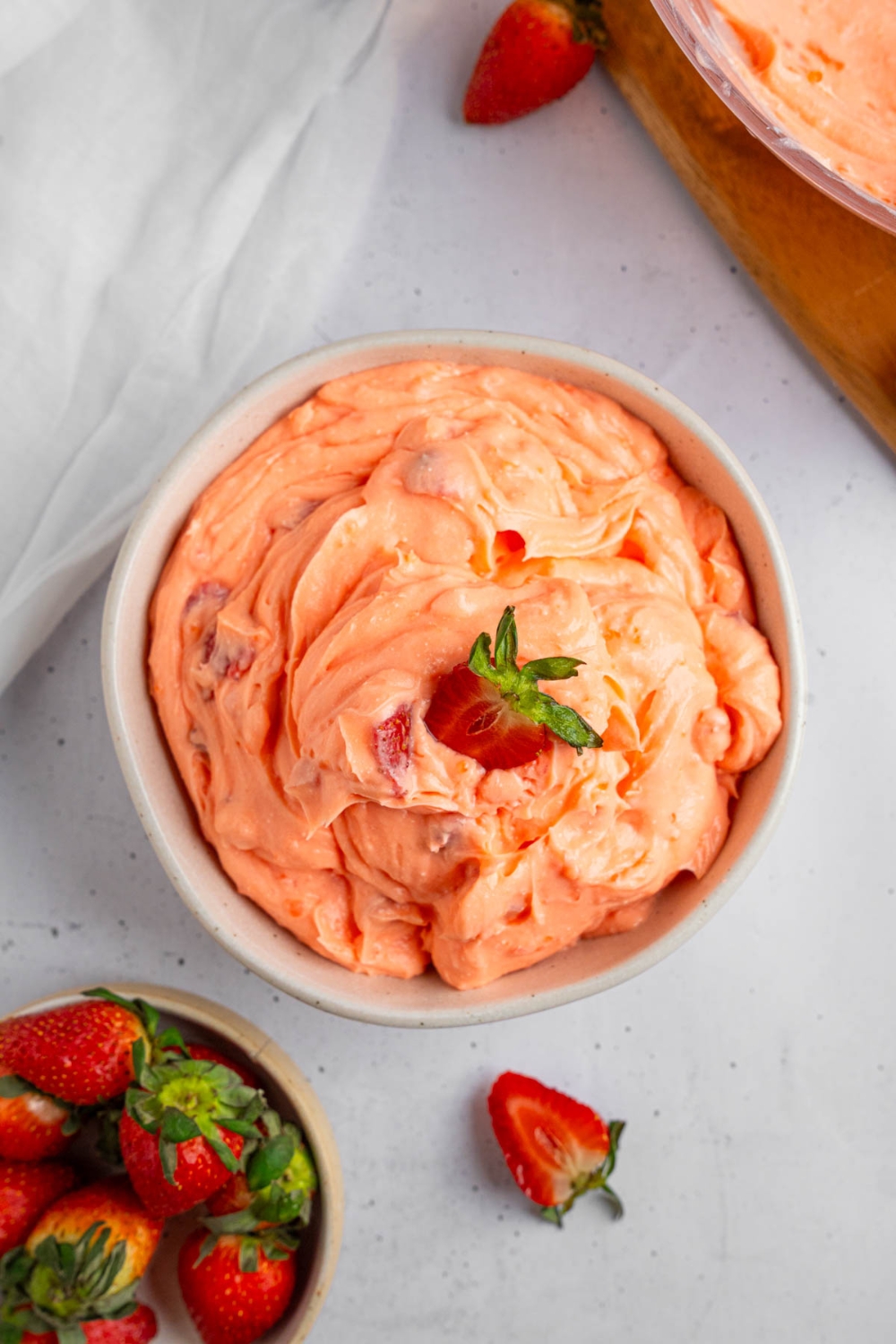 A white bowl of strawberry fluff salad with a sliced strawberry on top. The bowl is on a white counter with strawberries and a white cloth napkin.