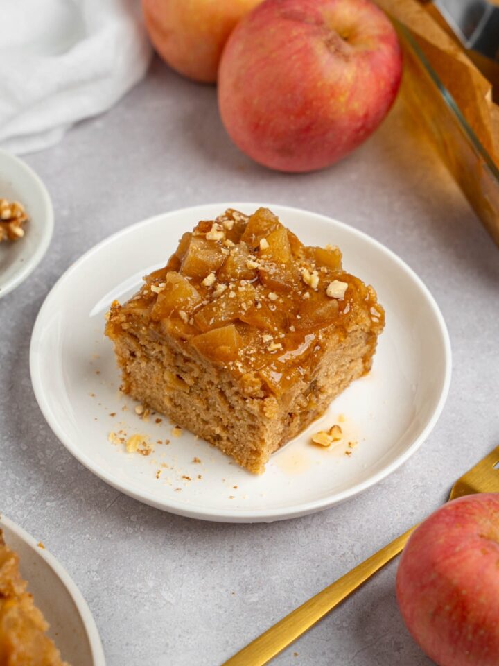 A slice of Amish applesauce cake on a white plate topped with chopped walnuts. The plate is on a white counter with apples and a fork.