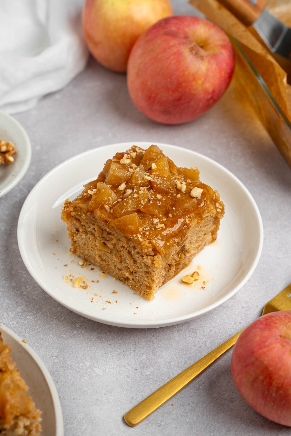 A slice of Amish applesauce cake on a white plate topped with chopped walnuts. The plate is on a white counter with apples and a fork.