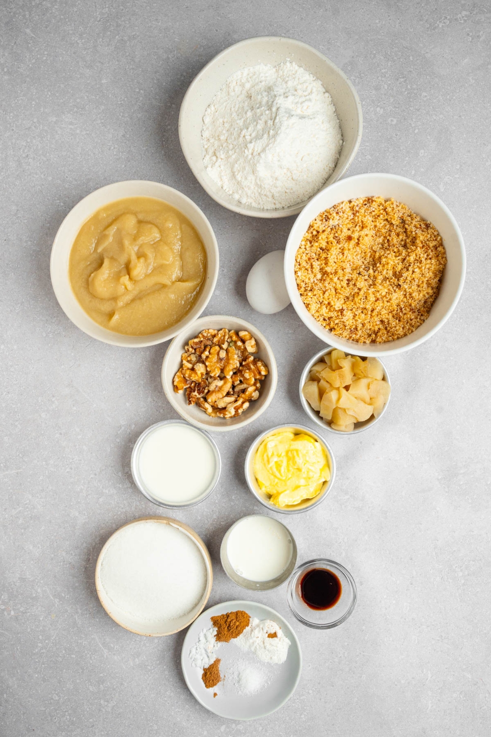 An overhead shot of several bowls in various sizes containing ingredients to make Amish applesauce cake including flour, baking soda, white sugar, brown sugar, milk, apples, butter, heavy cream, vanilla, and spices.
