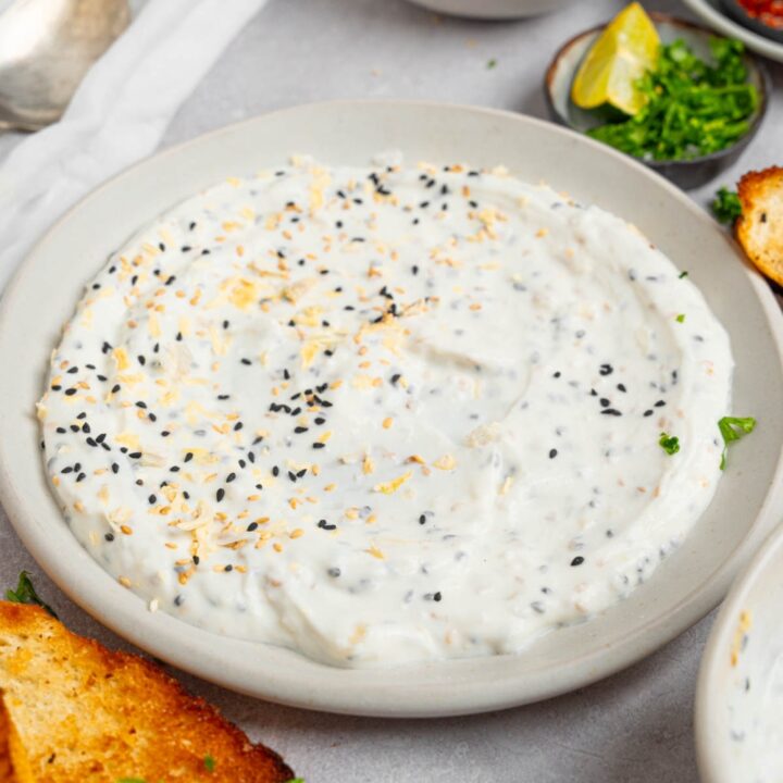 Bagel dip served in a white plate on a white counter with toasted bread and small ramekins of garnishes.