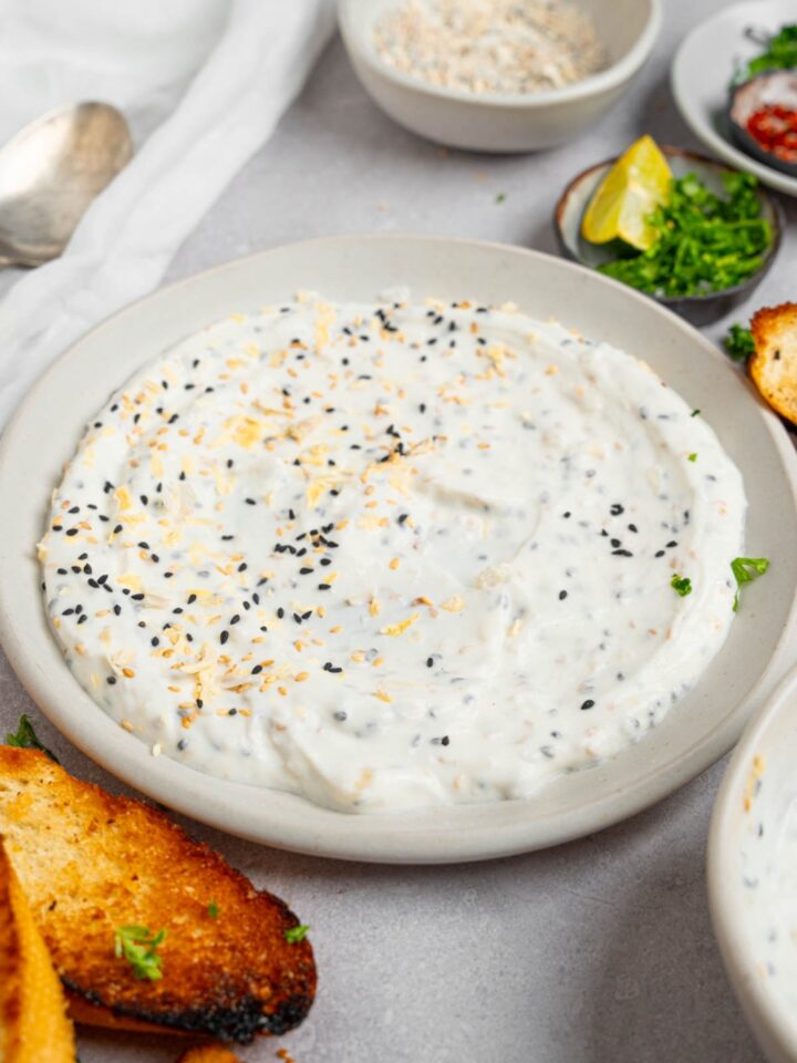 Bagel dip served in a white plate on a white counter with toasted bread and small ramekins of garnishes.