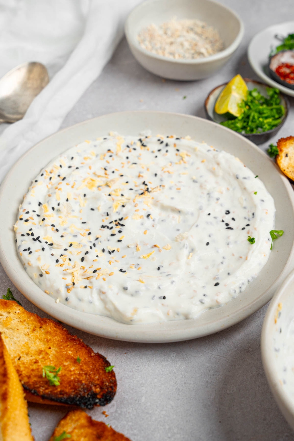 Bagel dip served in a white plate on a white counter with toasted bread and small ramekins of garnishes.