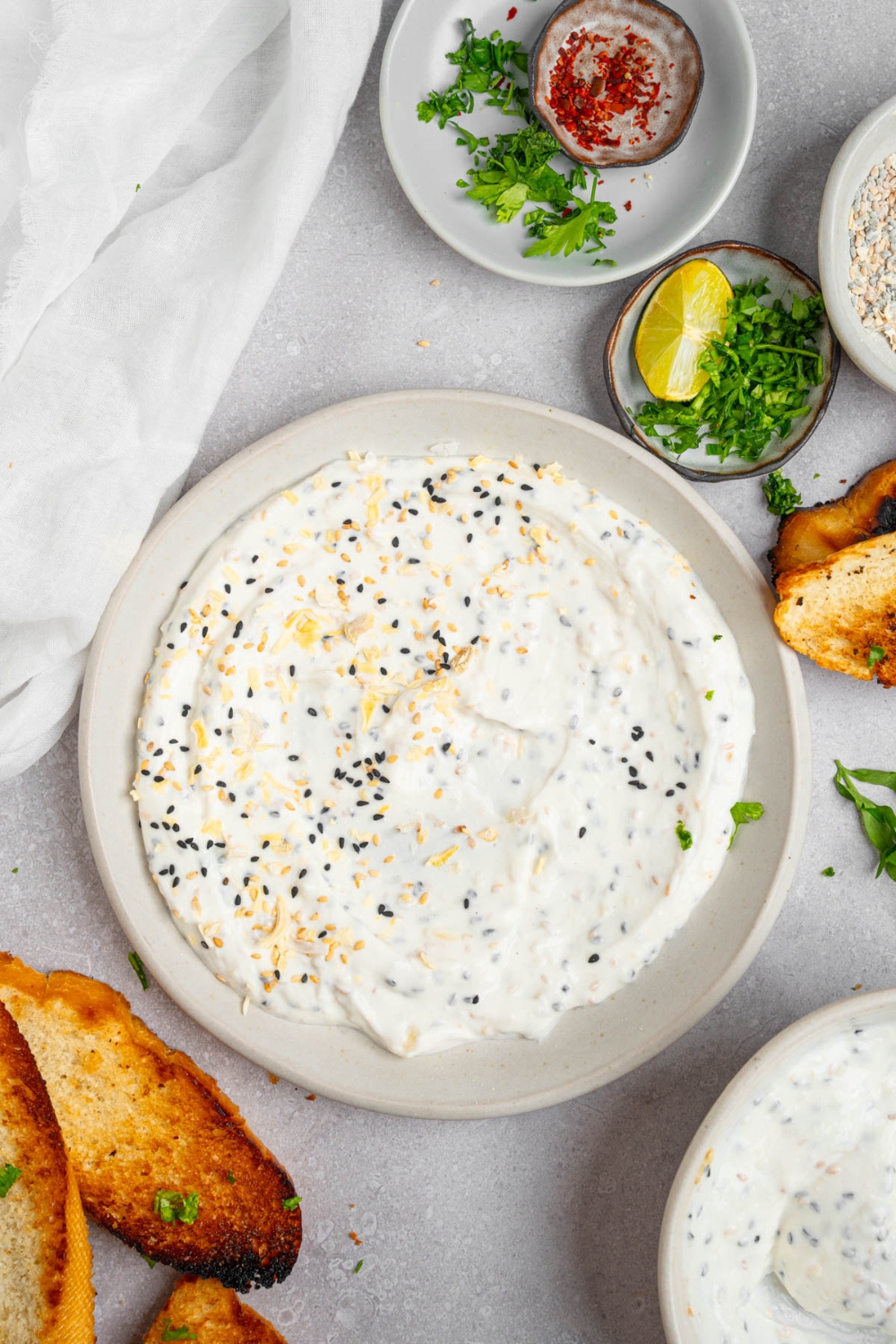 Bagel dip served in a white plate on a white counter with toasted bread and small ramekins of garnishes.