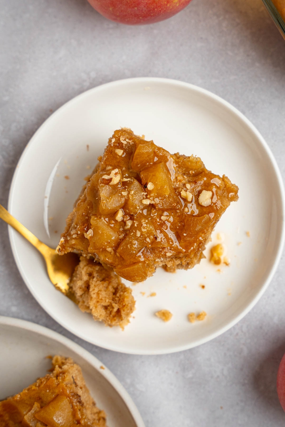 A slice of Amish applesauce cake topped with chopped walnuts on a white plate. A fork is on the plate with a bite of cake. The plate is on a white counter.