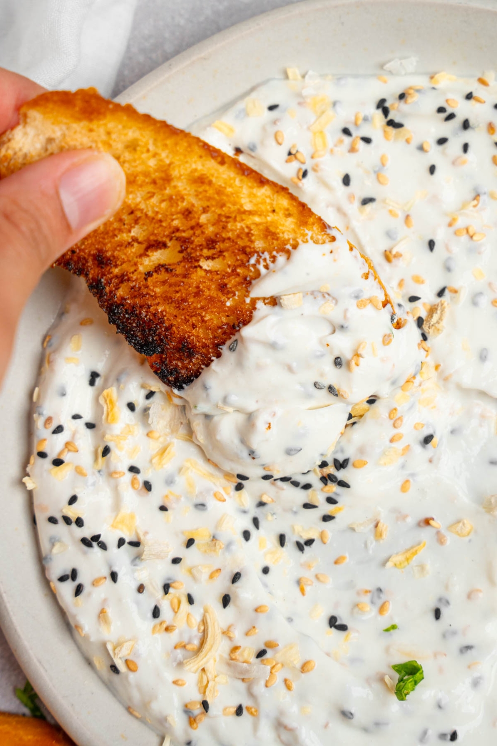 A hand dipping a slice of toasted bread in bagel dip served in a white plate.