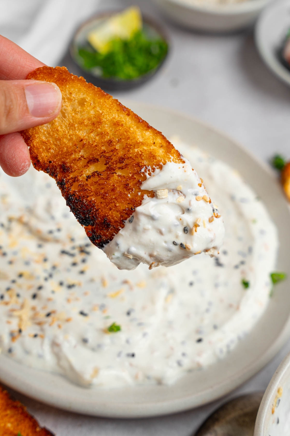 A close up of a hand holding a piece of toasted bread dipped in bagel dip. There is a plate of bagel dip on a white counter blurred in the background.