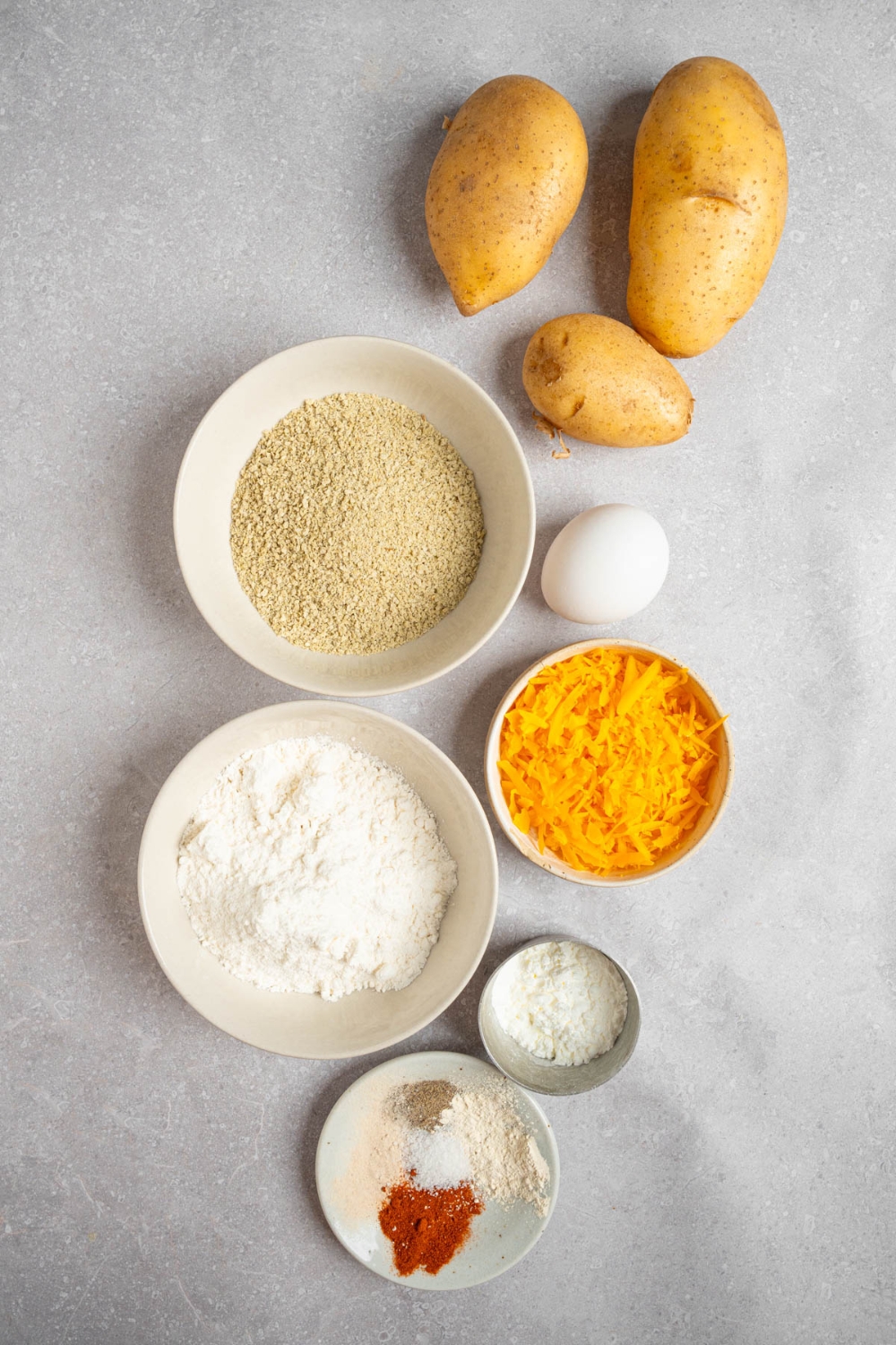 An overhead shot of several bowls in various sizes containing ingredients to make cheesy mashed potatoes including shredded cheese, potatoes, flour, egg, panko breading, and seasonings.
