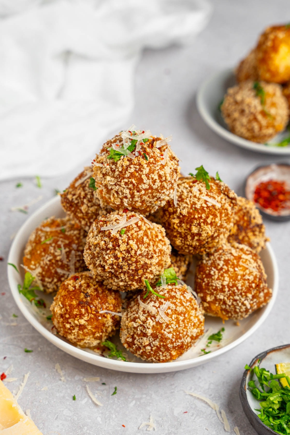 A pile of cheesy mashed potato balls served on a white plate garnished with fresh parsley and cheese. The plate is on a white counter with a white napkin and small ramekins of garnishes.