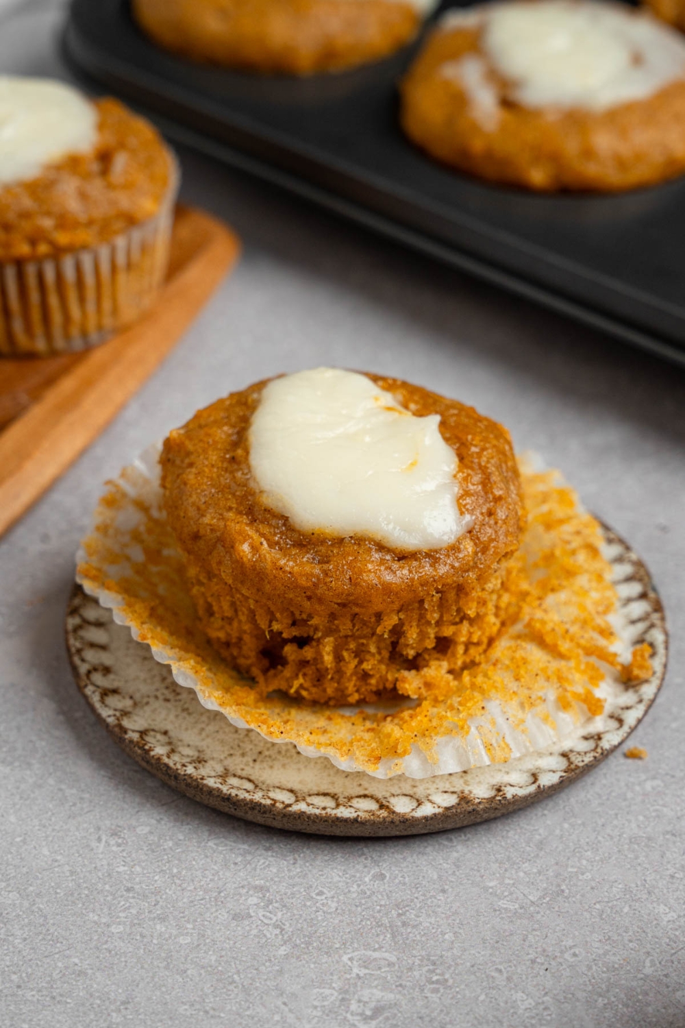An open homemade Starbucks pumpkin muffin topped with cream cheese filling on a small ceramic plate. The plate is on a white counter with a tray of baked pumpkin muffins.