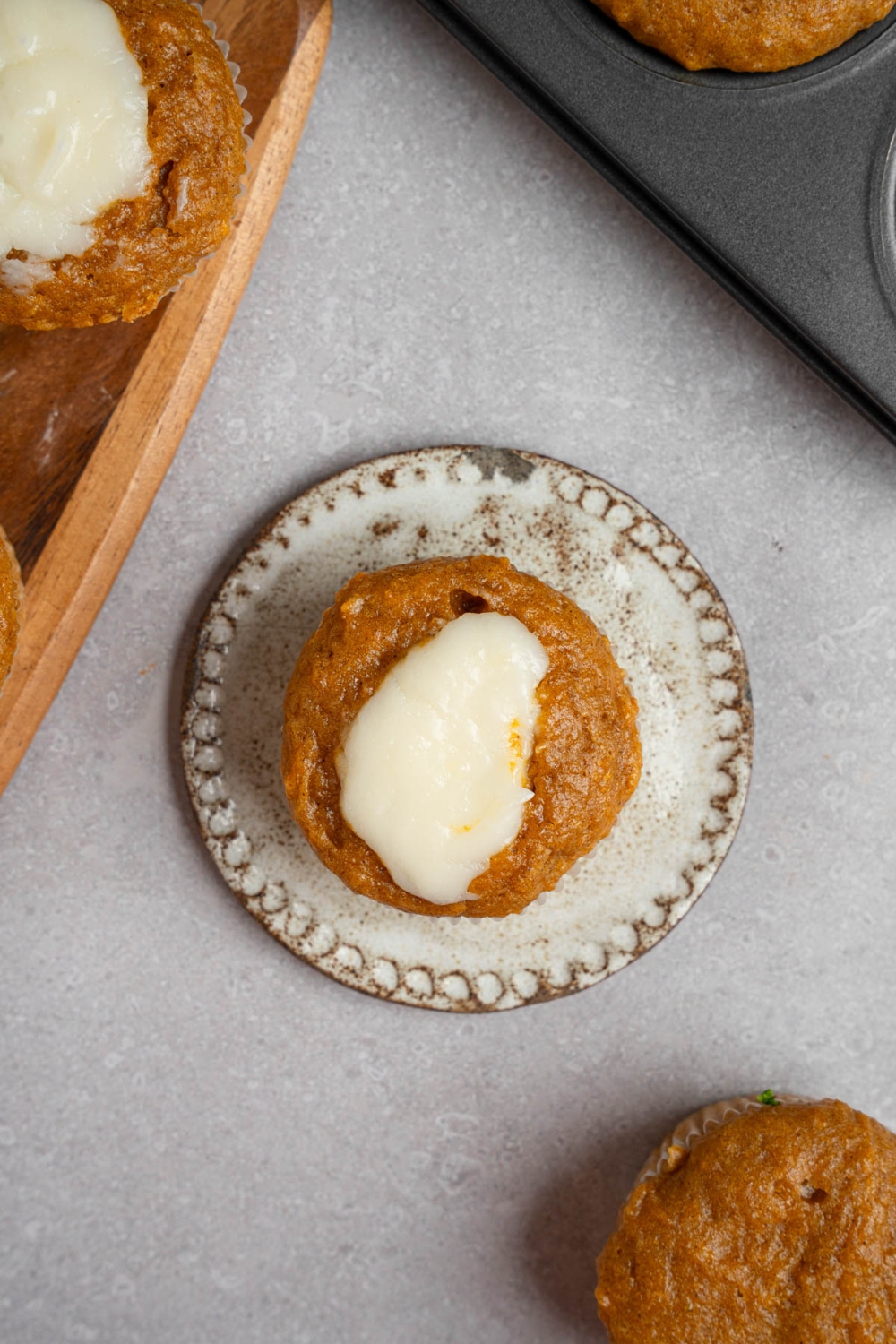 A homemade Starbucks pumpkin muffin topped with cream cheese filling on a small ceramic plate. The plate is on a white counter with a tray of baked pumpkin muffins.