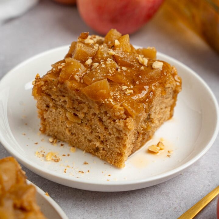 A slice of Amish applesauce cake on a white plate topped with chopped walnuts. The plate is on a white counter with apples and a fork.