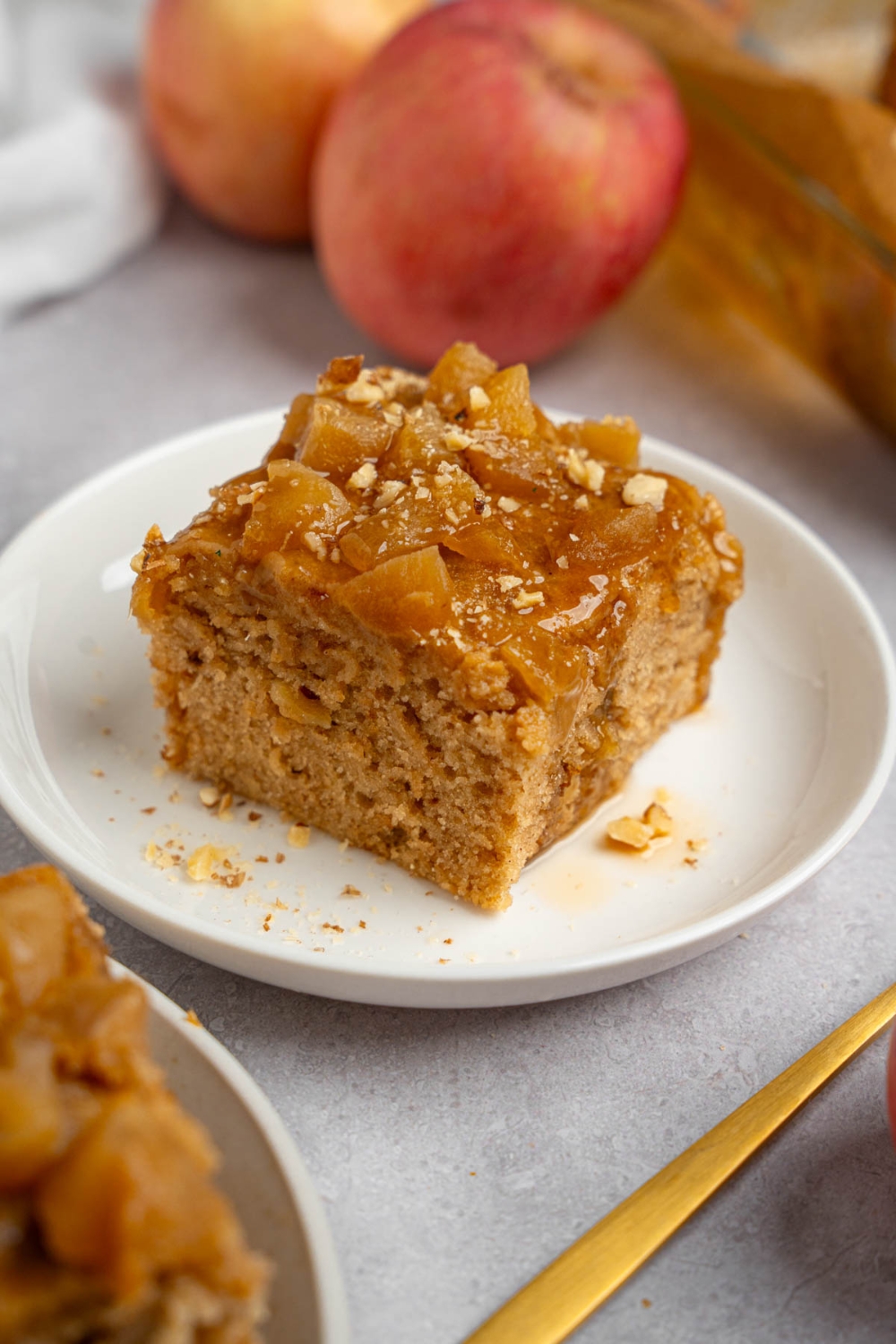 A slice of Amish applesauce cake on a white plate topped with chopped walnuts. The plate is on a white counter with apples and a fork.