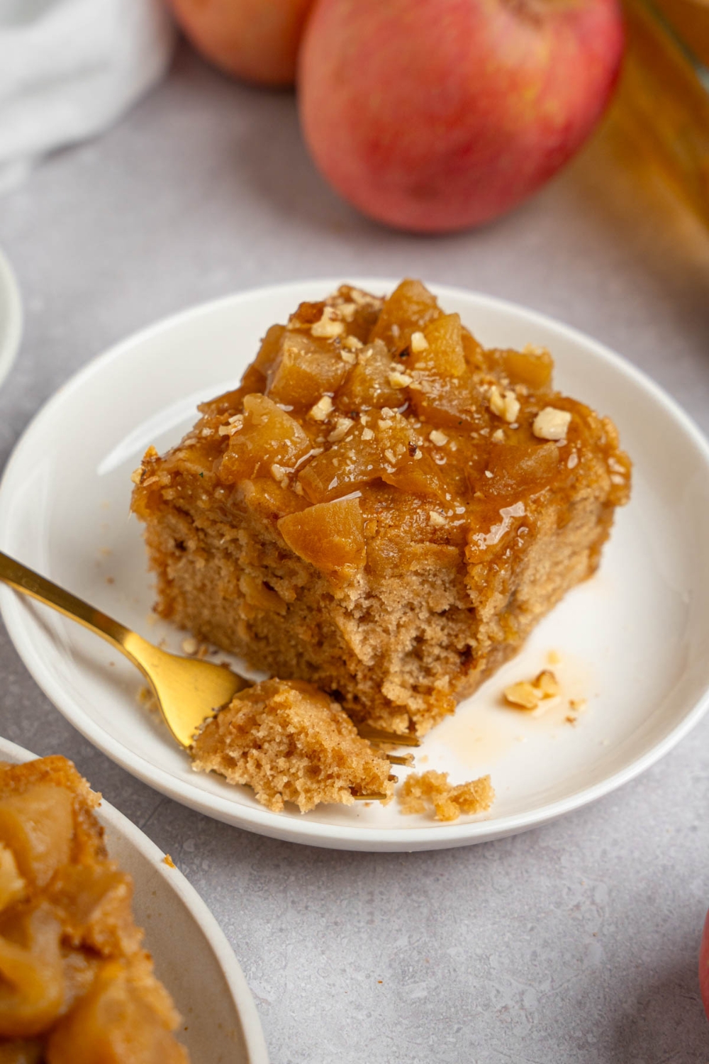 A slice of Amish applesauce cake topped with chopped walnuts on a white plate. A fork is on the plate with a bite of cake. The plate is on a white counter.