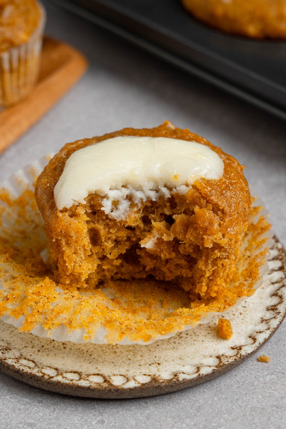 An open homemade Starbucks pumpkin muffin topped with cream cheese filling on a small ceramic plate. There is a bite taken from the muffin. The plate is on a white counter with a tray of baked pumpkin muffins.