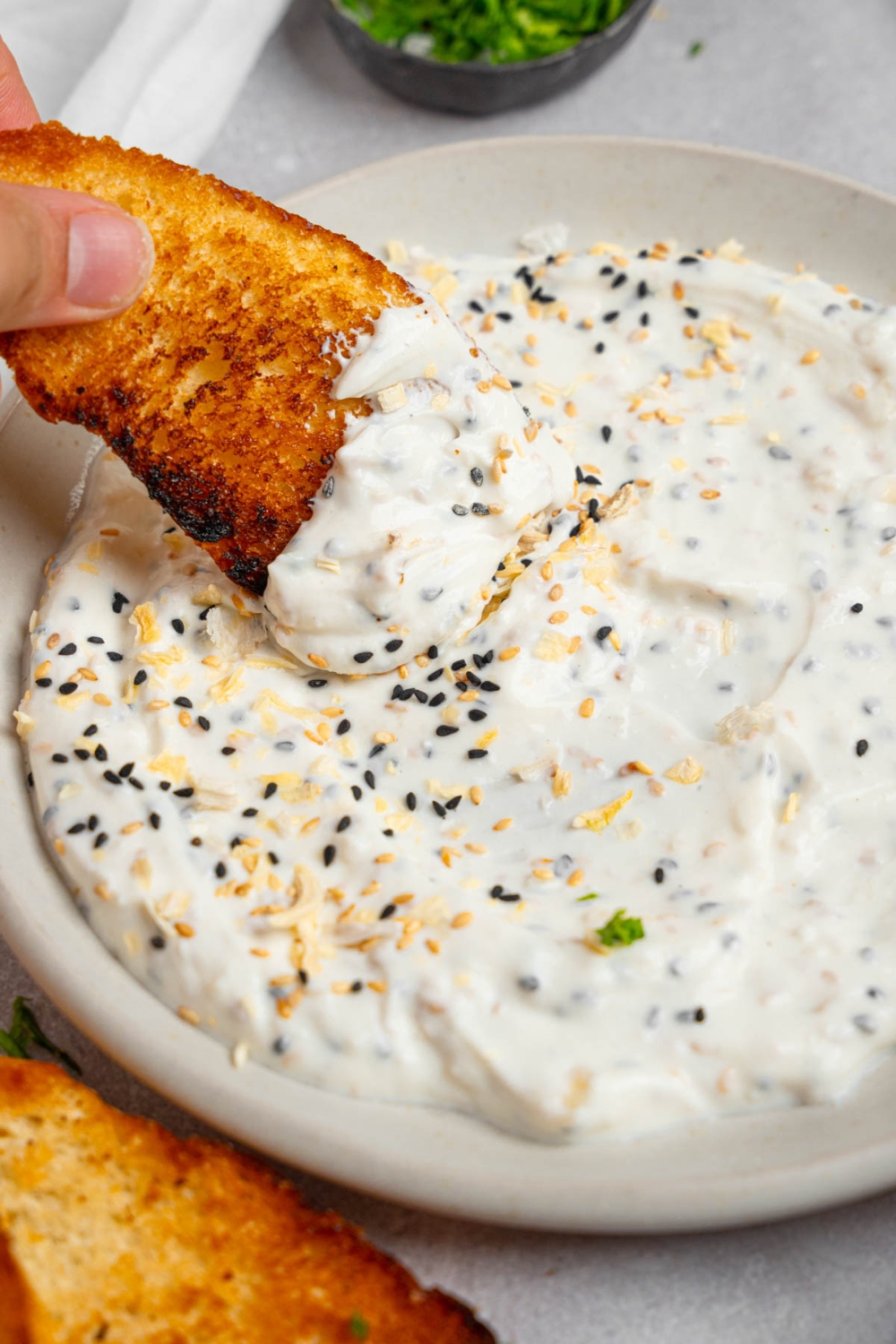A hand dipping a slice of toasted bread in bagel dip served in a white plate.