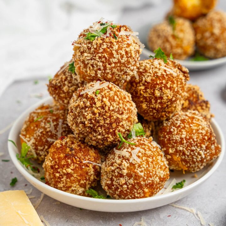A pile of cheesy mashed potato balls served on a white plate garnished with fresh parsley and cheese. The plate is on a white counter with an additional plate of mashed potato balls.