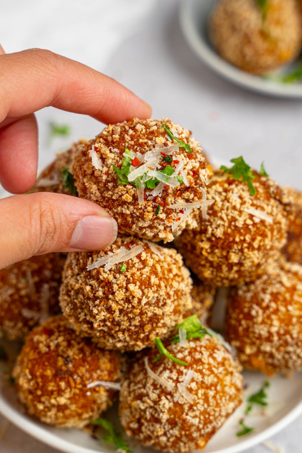 A pile of cheesy mashed potato balls served on a white plate garnished with fresh parsley and cheese. A hand is taking one of the mashed potato balls.