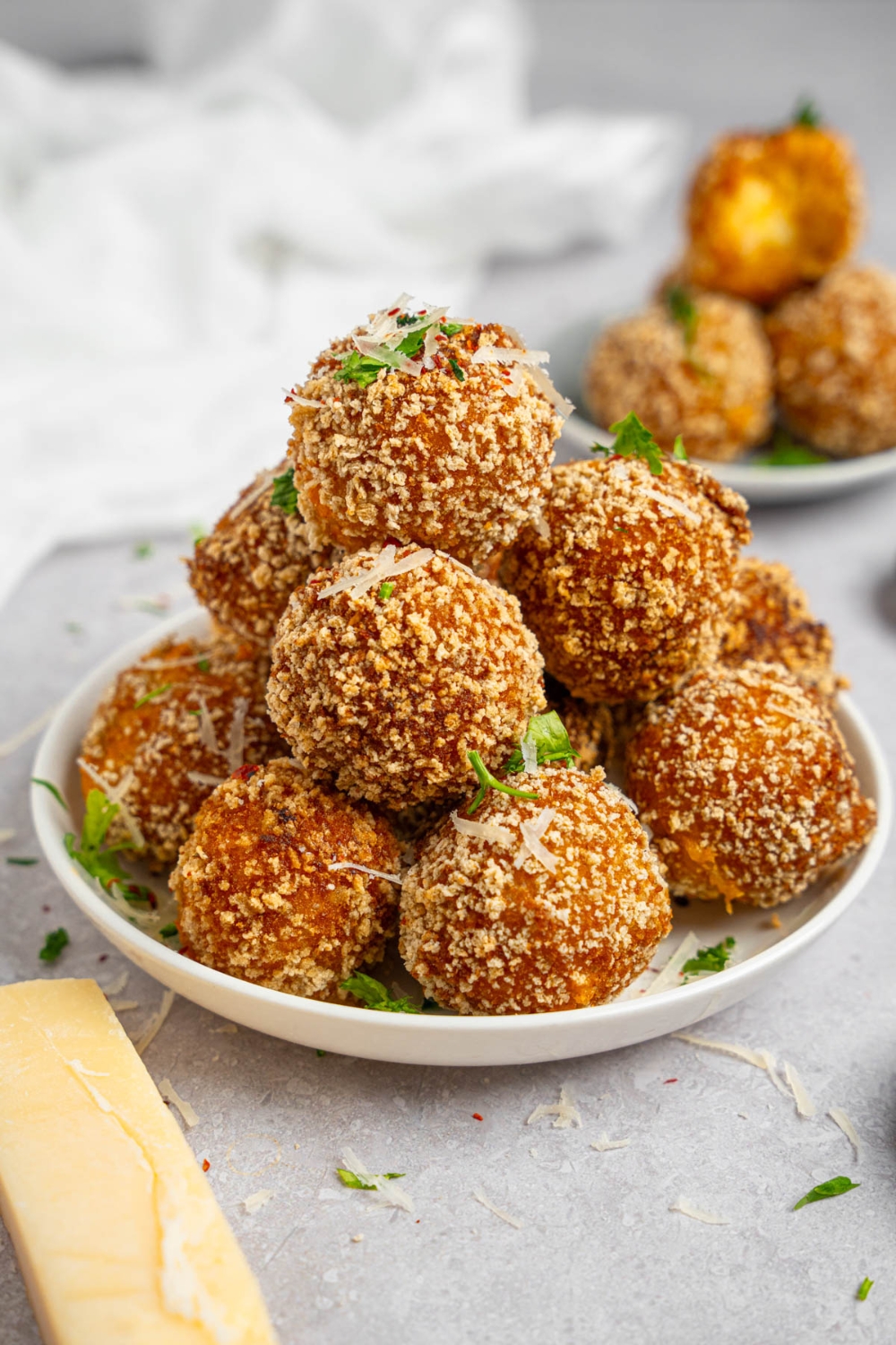 A pile of cheesy mashed potato balls served on a white plate garnished with fresh parsley and cheese. The plate is on a white counter with an additional plate of mashed potato balls.