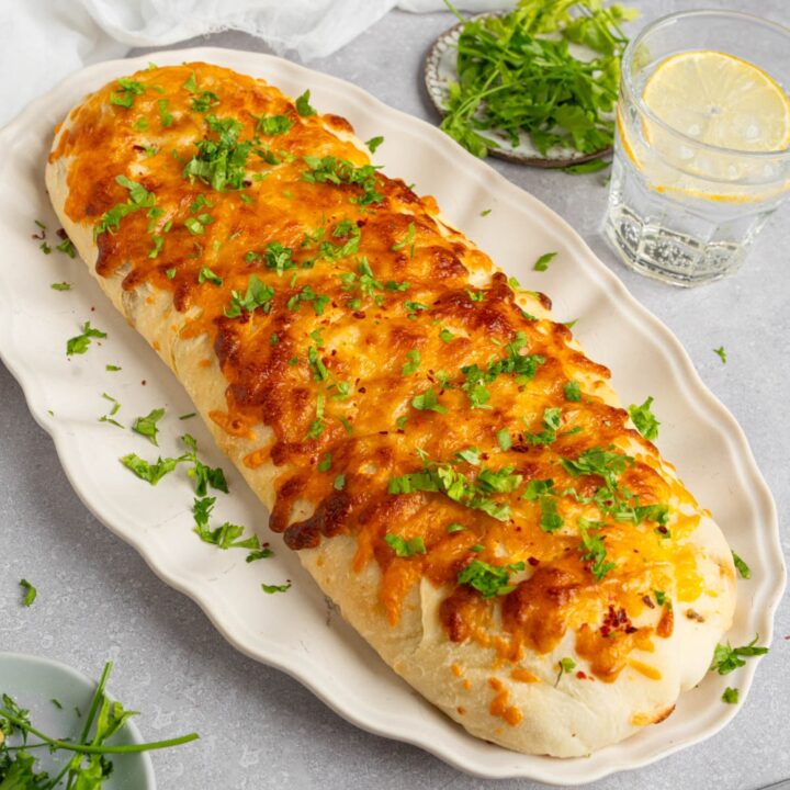 A loaf of garbage bread on a white platter garnished with chopped parsley. The platter is on a white counter with a glass of lemon water and small ramekins of garnishes.
