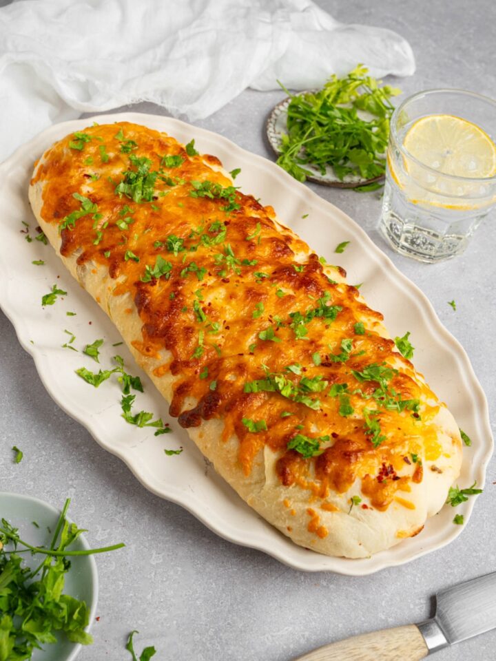 A loaf of garbage bread on a white platter garnished with chopped parsley. The platter is on a white counter with a glass of lemon water and small ramekins of garnishes.