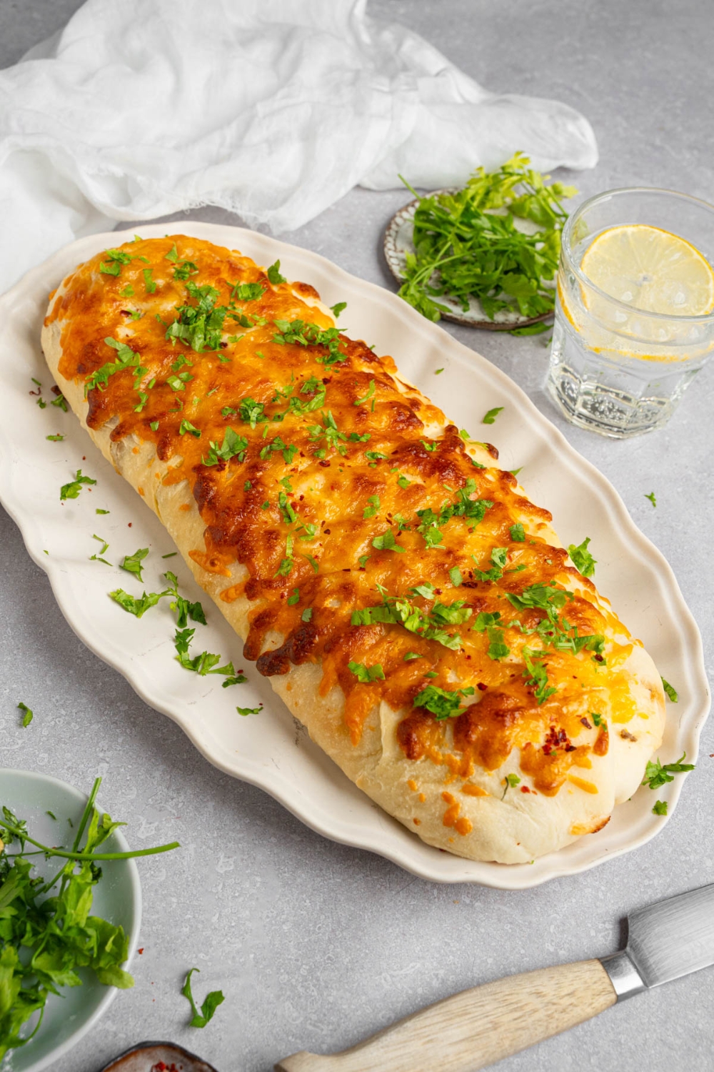 A loaf of garbage bread on a white platter garnished with chopped parsley. The platter is on a white counter with a glass of lemon water and small ramekins of garnishes.