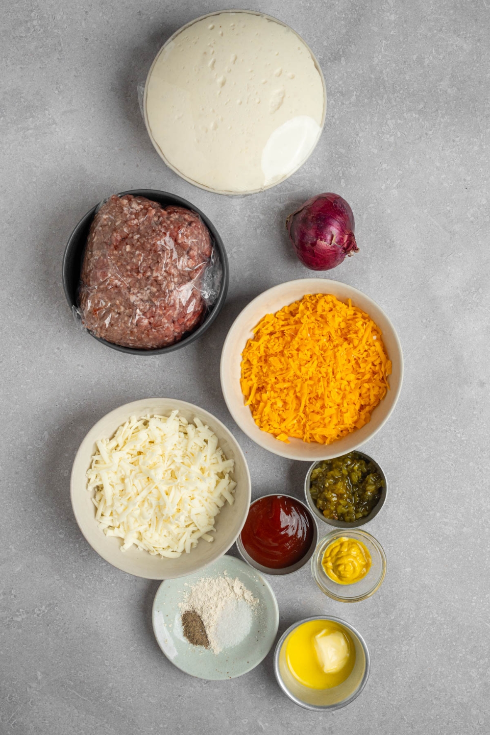 An overhead shot of several bowls in various sizes containing ingredients to make garbage bread including ground beef, pizza dough, shredded cheddar cheese, red onion, shredded pepper jack cheese, ketchup, mustard, relish, and seasonings.
